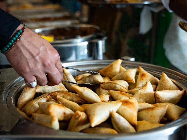 A hand holding metal tongs is picking samosas from a large silver dish filled with golden-brown, crispy samosas at a wedding catered by The Clay Oven. The person is wearing a beaded bracelet and a ring. In the background, there are stainless steel containers with more food items, slightly out of focus.
