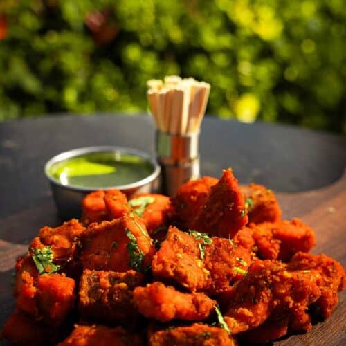 A wooden platter topped with pieces of spicy, deep-fried chicken seasoned with red spices and garnished with chopped herbs. In the background, a small metal cup filled with green dipping sauce and a container holding wooden toothpicks sit against a leafy garden setting—perfect for an exquisite wedding venue.