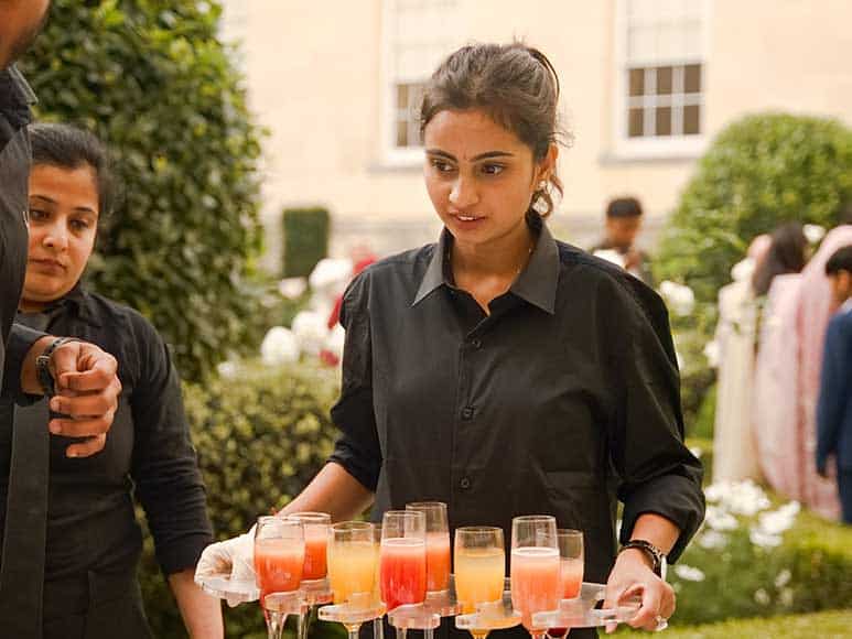 A woman wearing a black uniform, catering for The Clay Oven, carries a tray with multiple glasses containing colorful beverages. She stands outside in a garden area near neatly trimmed bushes. Another person in black is visible in the background, along with several blurred figures and a beige building facade.